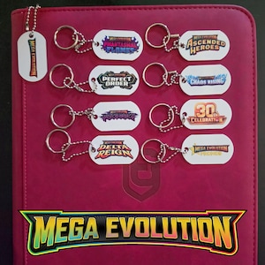 May include: A collection of nine white dog tag keychains with metal ball chains and split rings. Each tag features colorful graphic logos with text including Mega Evolution, Phantasmal Flames, Ascended Heroes, Perfect Order, Chaos Rising, Pitch Black, 30th Celebration, Delta Reign, and Promos.