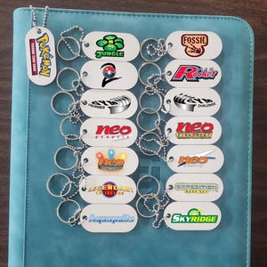 Może przedstawiać: Kolekcja białych metalowych breloków z logo i tekstem z gry karcianej Pokémon. Breloki zawierają logo "Jungle", "Fossil", "Rocket", "Neo Genesis" i "Aquapolis". Breloki są ułożone na niebieskiej powierzchni.