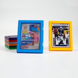 May include: A blue and yellow card holder with a stack of other colored holders. The blue holder displays a Pokemon card with a pink creature. The yellow holder displays a football card. The cards are protected by clear plastic.