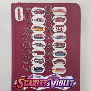 May include: A collection of white rectangular keychains with rounded corners, each featuring a unique Scarlet & Violet logo. The keychains are arranged in two columns on a dark pink surface, with a large Scarlet & Violet logo at the bottom.