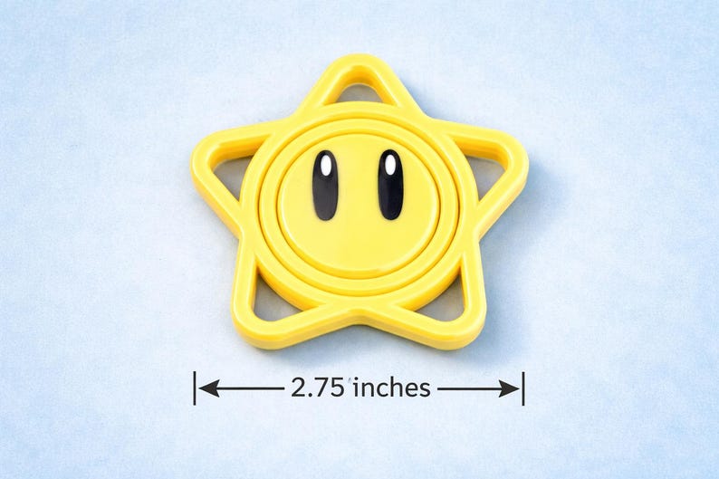 May include: A yellow star-shaped toy with a smiling face and black eyes. The toy measures 2.75 inches across. The star has a circular center and a star-shaped outline.
