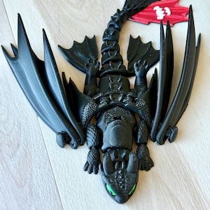 May include: A black dragon figurine with large wings and green eyes. The dragon is lying down with its wings spread. The figurine is made of a 3D-printed material and has a detailed design. The dragon's tail is curved, and the wings are large and pointed.