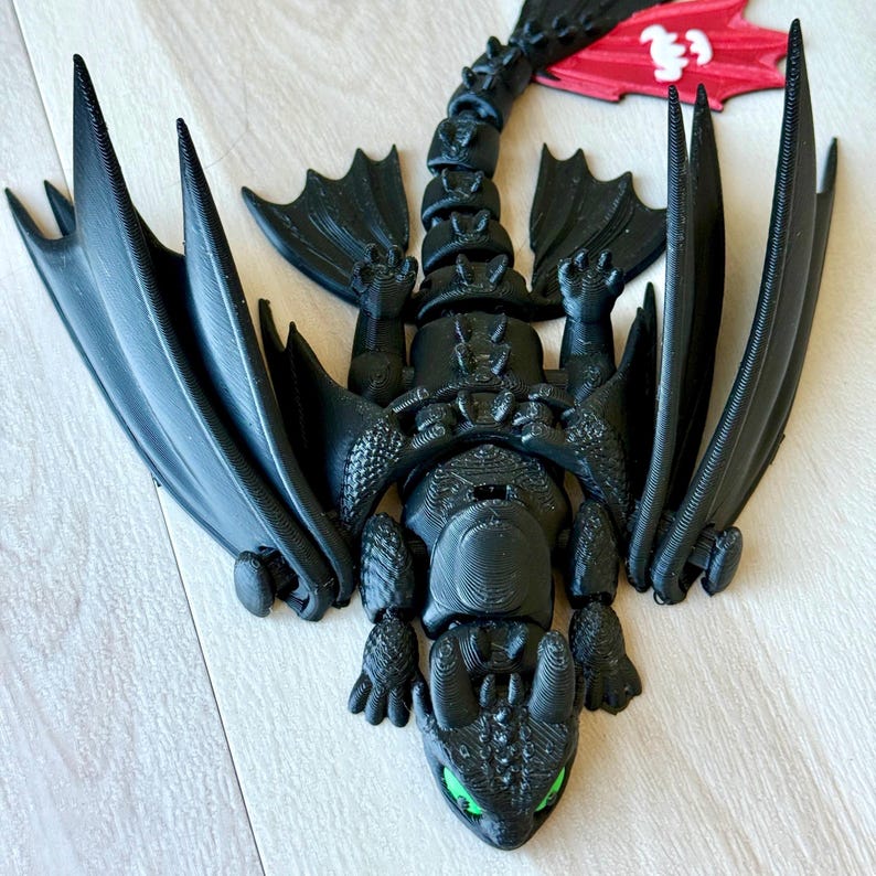 May include: A black dragon figurine with large wings and green eyes. The dragon is lying down with its wings spread. The figurine is made of a 3D-printed material and has a detailed design. The dragon's tail is curved, and the wings are large and pointed.