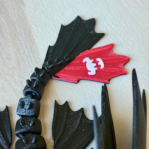 May include: A 3D-printed black dragon tail with articulated segments and wing-like fins. A red fin features a white emblem. The design is likely inspired by a popular animated film, showcasing a detailed and textured aesthetic.
