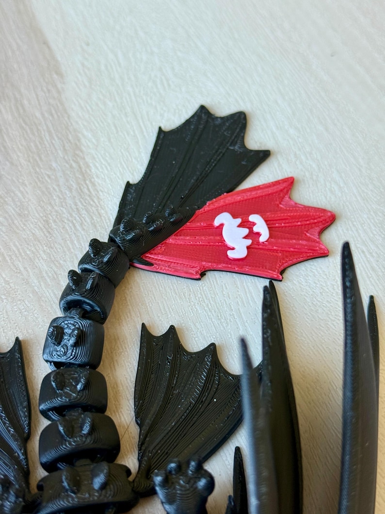 May include: A 3D-printed black dragon tail with articulated segments and wing-like fins. A red fin features a white emblem. The design is likely inspired by a popular animated film, showcasing a detailed and textured aesthetic.