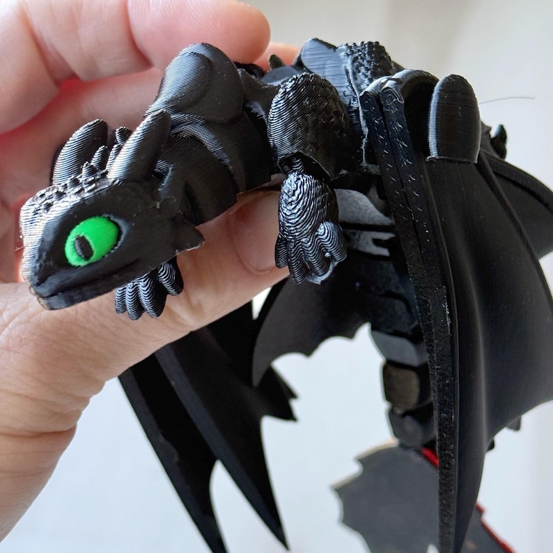 May include: A black dragon figurine with green eyes and large black wings. The dragon is a detailed 3D-printed model, featuring textured surfaces and intricate designs. The figurine is held in a hand.