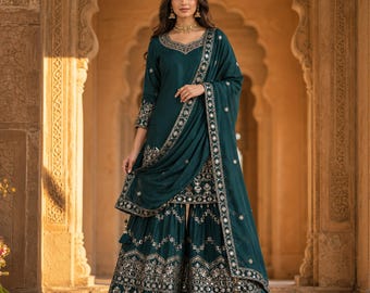 Puede incluir: Un conjunto sharara de color verde azulado oscuro que incluye una túnica de manga larga y pantalones anchos, adornados con intrincados bordados plateados y espejos. El conjunto incluye una dupatta a juego con un borde decorativo.