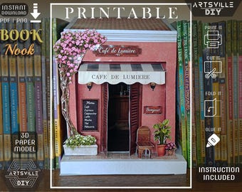 May include: A 3D paper model book nook featuring a pink cafe facade named Cafe de Lumiere. The structure includes a striped awning, a menu board, a small chair, a potted plant, and a climbing vine with pink flowers. Text includes Cafe de Lumiere, Menu, and Bonjour.