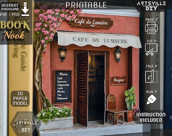May include: A 3D paper model book nook kit titled Café de Lumière, featuring a terracotta-colored building facade with a white awning, a wooden door, a menu board, a small chair, a potted plant, and a climbing vine with pink flowers.