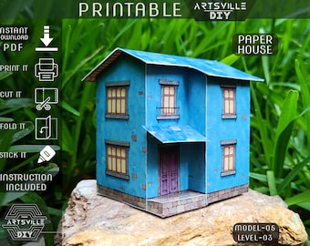 May include: A blue paper house model with a blue roof, windows, and a purple door. The model is labeled "Printable" and "Paper House". Instructions are included. The model is on a rock.