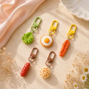May include: A set of five food-themed keychains featuring miniature charms of a green lettuce leaf, a fried egg, an orange carrot, a brown sausage, and a beige steamed bun, each attached to a matching coloured plastic lobster clasp hook.