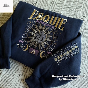 May include: Dark blue sweatshirt featuring gold embroidery. The design includes the word "ESQUIE", a sunburst, moons, and the text "EXPEDITION 33" and "2025". The sleeve has a matching design.