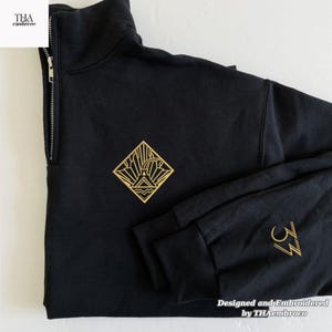 May include: Black quarter-zip sweatshirt with gold embroidered geometric design on the chest and sleeve. The design includes a stylized mountain and sun. The sleeve also features the number 33 in gold. The text "Designed and Embroidered by THAembroco" is at the bottom.