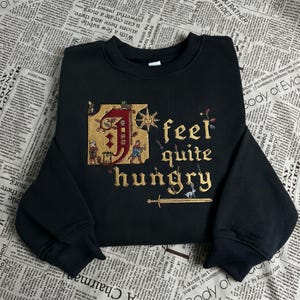 May include: Black sweatshirt with the phrase "feel quite hungry" in gold lettering. The design includes a medieval-style illustration with a large letter "J" and small figures. The sweatshirt has long sleeves.