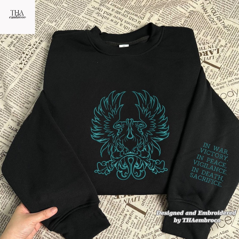 May include: Black sweatshirt with a teal embroidered emblem of a bird-like creature. The sleeve displays the text "IN WAR, VICTORY, IN PEACE, VIGILANCE, IN DEATH, SACRIFICE." Designed and embroidered by THAembroco, this sweatshirt is a unique apparel item.