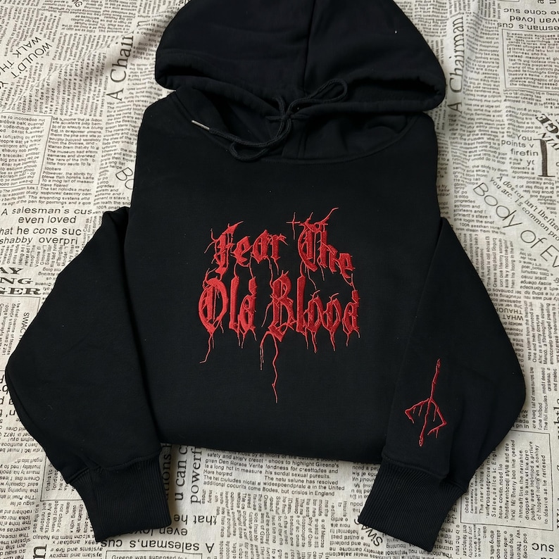 May include: Black hooded sweatshirt with the embroidered text "Fear The Old Blood" in red. The sweatshirt also features a red embroidered design on the left sleeve. The hoodie has a drawstring and a hood.