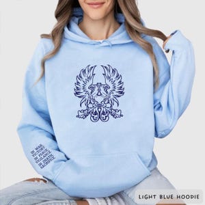 May include: Pale blue hoodie featuring a dark blue winged emblem. The hoodie has a front pocket and a drawstring hood. Text on the sleeve reads "IN WAR, VICTORY. IN PEACE, VIGILANCE. IN DEATH, SACRIFICE."