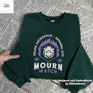 Può includere: Felpa verde scuro con la scritta ricamata "GRAND NECROPOLIS - NEVARRA CITY" e "MOURN WATCH" in bianco e viola. Il design include un teschio e una grafica a fiamma. Progettato e ricamato da THAembroco.