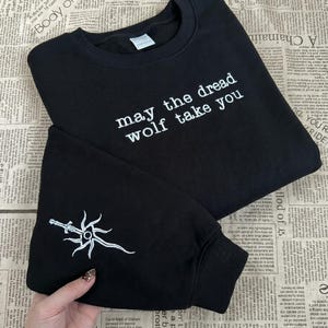May include: Black sweatshirt with the phrase "may the dread wolf take you" in white text. The left sleeve has a white sword and sun emblem. The garment is shown on a newspaper background.