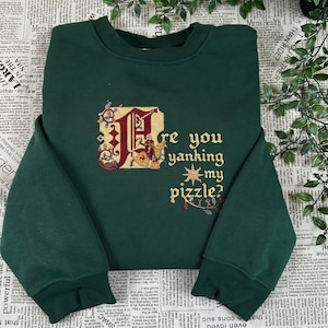 May include: A dark green crewneck sweatshirt featuring an embroidered medieval-style illuminated letter design. The embroidery includes a knight on horseback and the text "Are you yanking my pizzle?" in a gold-coloured gothic font with a sun icon.