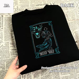 May include: Black sweatshirt with a teal and white embroidered design. The design features a wolf-like creature and a figure with the text "DREAD WOLF". The sweatshirt is designed and embroidered by THAembroco.