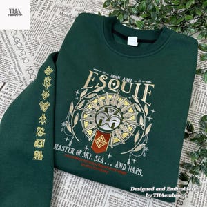 May include: A dark green sweatshirt with gold embroidery. The design includes the word "ESQUIF", a sunburst, and the text "MASTER OF SKY, SEA... AND NAPS." The sleeve has additional gold embroidered symbols. Designed and embroidered by THAembroco.