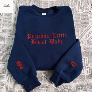 May include: Navy blue sweatshirt with the text "Precious Little Bhaal Babe" embroidered in red gothic lettering. Features a red graphic on each sleeve. A comfortable, casual top.