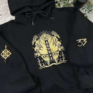 Puede incluir: Sudadera con capucha negra con bordado dorado. La parte delantera presenta una figura estilizada con los números "20", "25" y "37". Un diseño geométrico dorado adorna la manga izquierda. La sudadera tiene una capucha con cordón.