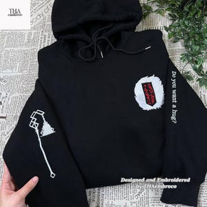 May include: Black hoodie with white embroidered details. Features a design on the left sleeve and a crest on the chest. The right sleeve has the text "Do you want a hug?" The hoodie is designed and embroidered by THAembroco.