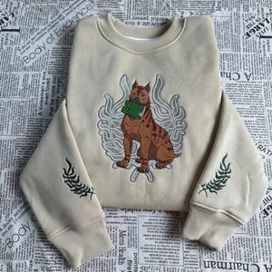 Puede incluir: Sudadera de color crema con un perro marrón bordado sosteniendo un libro verde. El diseño está rodeado por una corona decorativa. Bordados verdes en forma de helecho adornan las mangas. Una prenda casual y cómoda.