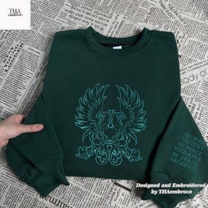 May include: A dark green sweatshirt featuring a teal emblem of a bird or mythical creature. The sleeve displays embroidered text: "IN WAR, VICTORY, IN PEACE, VIGILANCE, IN DEATH, SACRIFICE". Designed and embroidered by THAembroco. The sweatshirt is a casual garment.