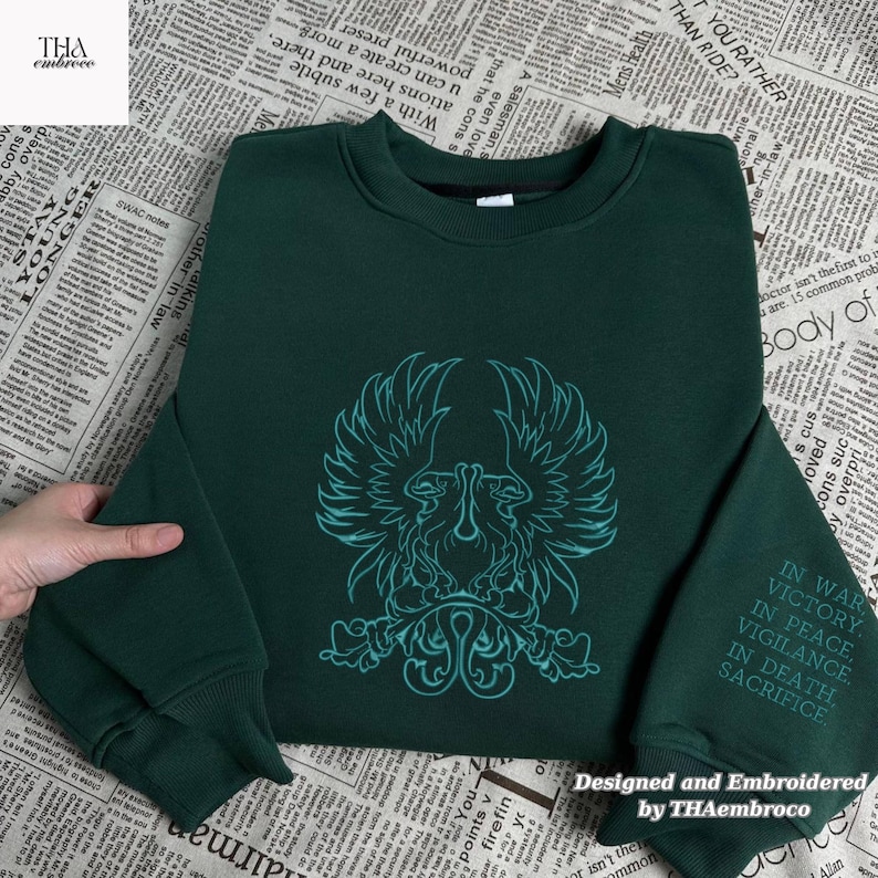 May include: A dark green sweatshirt featuring a teal emblem of a bird or mythical creature. The sleeve displays embroidered text: "IN WAR, VICTORY, IN PEACE, VIGILANCE, IN DEATH, SACRIFICE". Designed and embroidered by THAembroco. The sweatshirt is a casual garment.