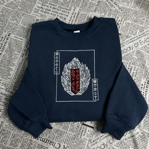 Puede incluir: Sudadera azul marino con un diseño gráfico blanco y rojo. El diseño presenta una forma estilizada de llama o ala con patrones geométricos y símbolos dentro de un marco rectangular blanco. Se muestra sobre un fondo de periódico.