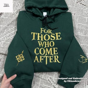 Może przedstawiać: Ciemnozielona bluza z kapturem ze złotym haftowanym tekstem „FOR THOSE WHO COME AFTER”. Bluza z kapturem ma kaptur ze sznurkiem i złote haftowane wzory na rękawach. Widoczny jest również tekst „Designed and Embroidered by THAembroco”.