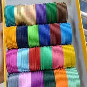 May include: A box filled with stacks of colorful bangles. The bangles are in various colors, including gold, pink, purple, green, and orange. The box is yellow, and the bangles are neatly arranged in rows.