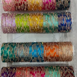May include: Four stacks of colourful glass bangles in a clear plastic container. The bangles are in shades of pink, red, yellow, green, blue, and orange, with gold-coloured accents. The bangles are likely for a woman.