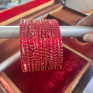 Könnte beinhalten: Ein Set roter Armreifen mit goldfarbenen Akzenten. Die Armreifen werden auf einer silbernen Stange präsentiert, vor dem Hintergrund einer rot samtgefütterten Box. Die Armreifen bestehen aus kleinen, quadratisch geschliffenen roten Steinen.
