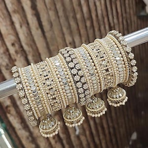 May include: A collection of ornate, gold-toned bangles with intricate detailing. The bangles are adorned with small, reflective embellishments and faux pearls. Each bangle features a dangling, bell-shaped charm, adding to the overall elegance.