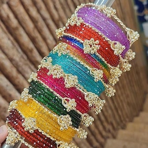 May include: A selection of vibrant bangles in a rainbow of colours, including purple, red, blue, green, and yellow. Each bangle is decorated with gold-coloured floral details. The bangles are displayed on a silver rod, highlighting their bright colours and intricate patterns.