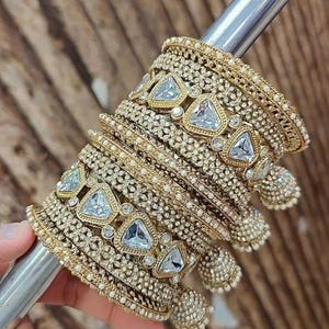 May include: A collection of gold-toned bangles with intricate detailing and clear, triangular gemstones. The bangles are stacked together, showcasing a variety of textures and designs, including small pearl-like accents. The jewelry is displayed on a silver rod.