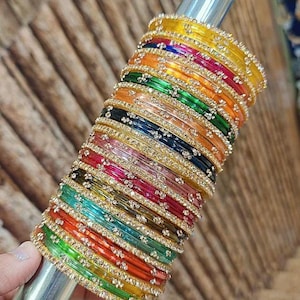 Puede incluir: Una colección de coloridas pulseras en varios tonos, incluyendo amarillo, naranja, verde y azul, se apilan en una varilla plateada. Cada pulsera está adornada con pequeños adornos brillantes, creando una exhibición vibrante y llamativa.