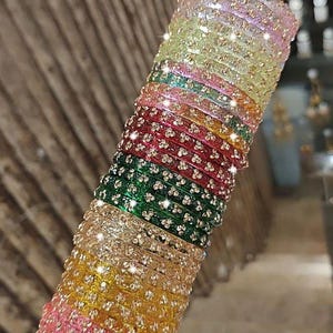 May include: A collection of multi-coloured bangles, featuring pink, yellow, green, and red hues, embellished with shimmering details. The bangles are arranged on a silver rod, creating a striking visual. These bangles are likely for special events.