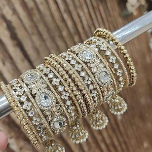 May include: A selection of gold-coloured bangles with detailed patterns, including floral and geometric shapes, and clear, sparkling gemstones. Some bangles have dangling bell-shaped charms with small pearls. The jewellery is displayed on a metal rod.