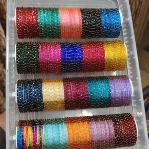 May include: A clear plastic container filled with colorful beaded bangles. The bangles are arranged in rows, showcasing a variety of colors including red, green, pink, blue, and gold. The bangles are likely for fashion or jewelry.