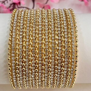 May include: A set of gold-toned bangles arranged in a stack. The set consists of alternating thin bands, featuring either a row of small, clear, reflective stones or a row of smooth, round, gold-coloured metal beads, creating a textured appearance.