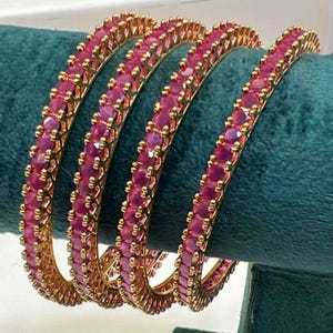 Puede incluir: Un conjunto de cuatro brazaletes circulares con una hilera continua de piedras redondas de color rosa vibrante, engastadas en un marco de metal dorado pulido. Cada brazalete presenta un diseño uniforme con engastes que crean un borde texturizado.