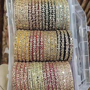 May include: A clear plastic container filled with stacks of colourful bangles. The bangles are decorated with small, reflective embellishments and come in various colours, including red, green, yellow, and pink.