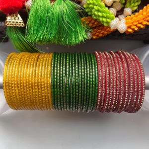 Puede incluir: Una colección de pulseras coloridas en amarillo, verde y rojo. Las pulseras amarillas tienen una superficie texturizada, mientras que las verdes y rojas son lisas. Las pulseras rojas están adornadas con pequeños adornos brillantes. Se ven borlas y guirnaldas decorativas en el fondo.