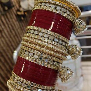 Puede incluir: Un conjunto de pulseras rojas y doradas con detalles intrincados. Las pulseras presentan una combinación de bandas rojas, adornos dorados y pequeños dijes colgantes en forma de campana. El diseño incorpora acentos espejados y cuentas perladas.
