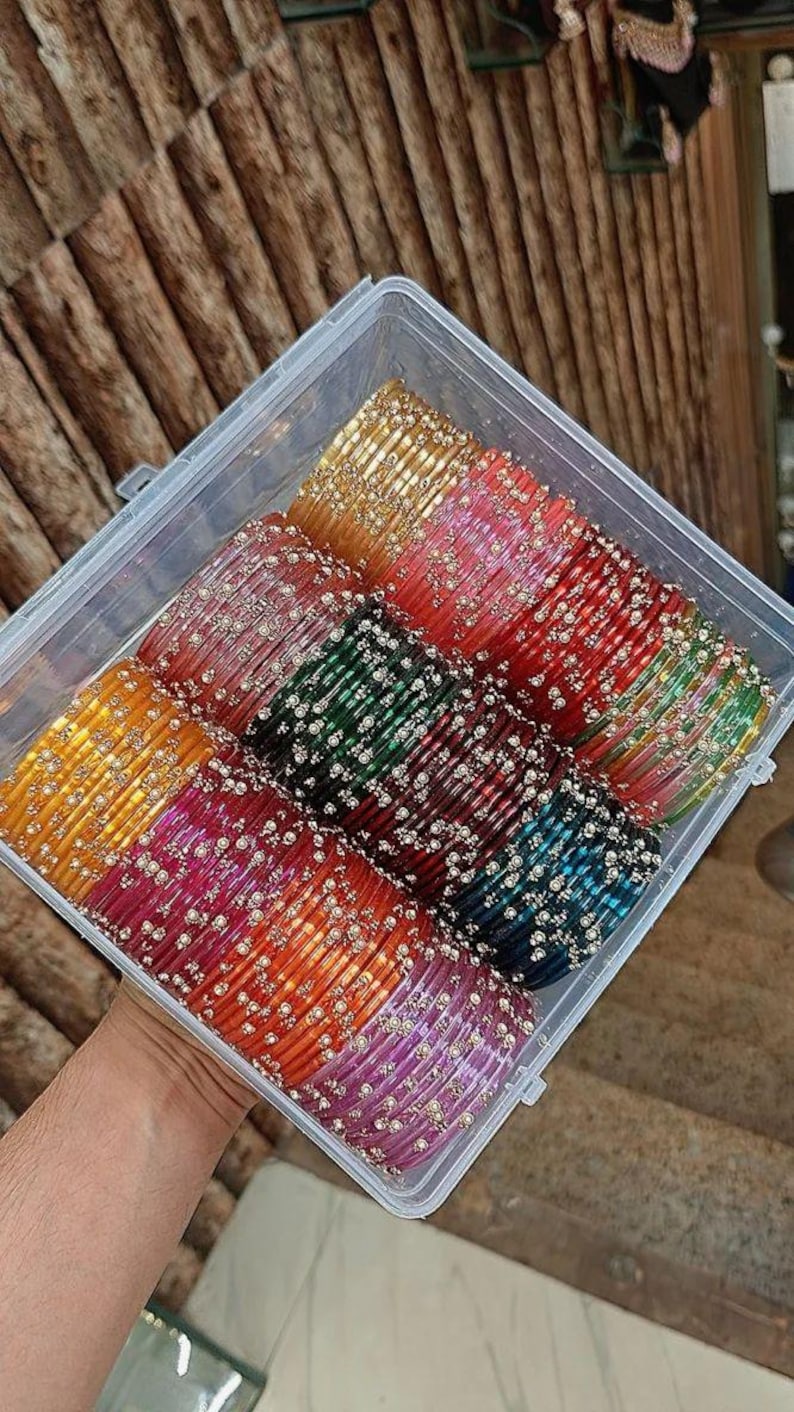 May include: A clear plastic container filled with colourful glass bangles. The bangles are in shades of gold, pink, green, red, and blue, and are decorated with small, silver-coloured beads. The bangles are stacked in rows.